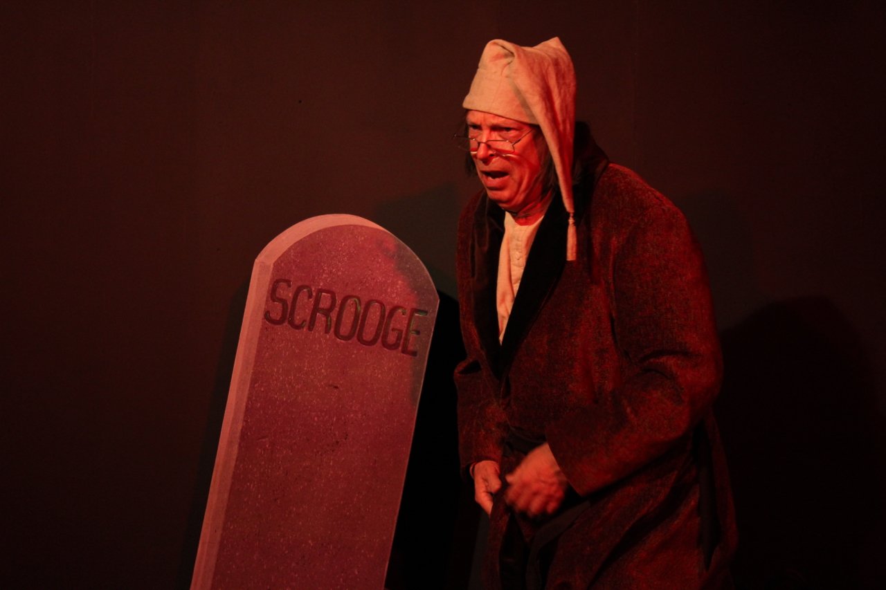 Actor portraying Scrooge in front of a tombstone labeled 'Scrooge' in a theatrical scene.