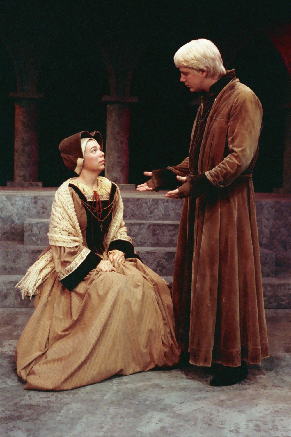 Two actors in medieval costumes performing a scene on stage, with a woman sitting and a man standing, possibly engaging in a dialogue.