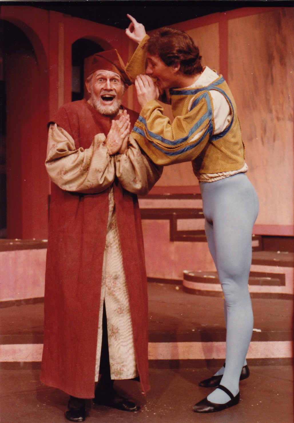 Two actors on stage dressed in medieval-style costumes, one in a long robe and hat, the other in a colorful tunic and tights, engaged in an animated conversation.