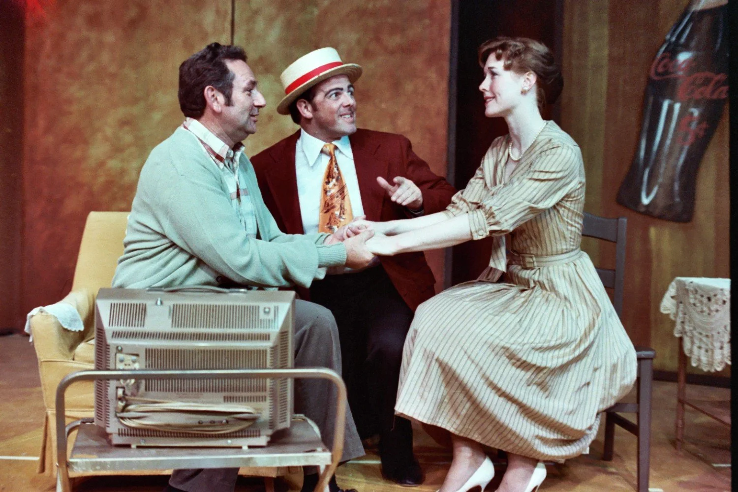 Three actors in a theatrical scene, the man on the left wearing a sweater and plaid shirt, the man in the middle wearing a red blazer and straw hat, and the woman on the right in a striped dress, holding hands and smiling. A vintage television set is