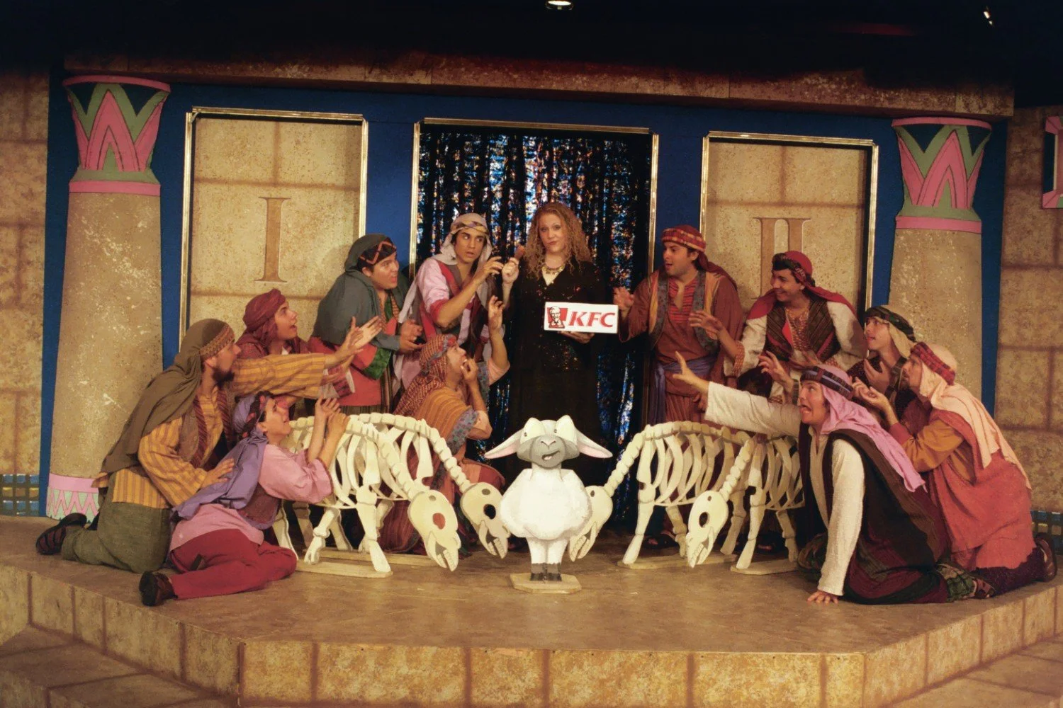 Stage performance with people in costumes surrounding a woman holding a KFC bucket, featuring sheep skeleton props.