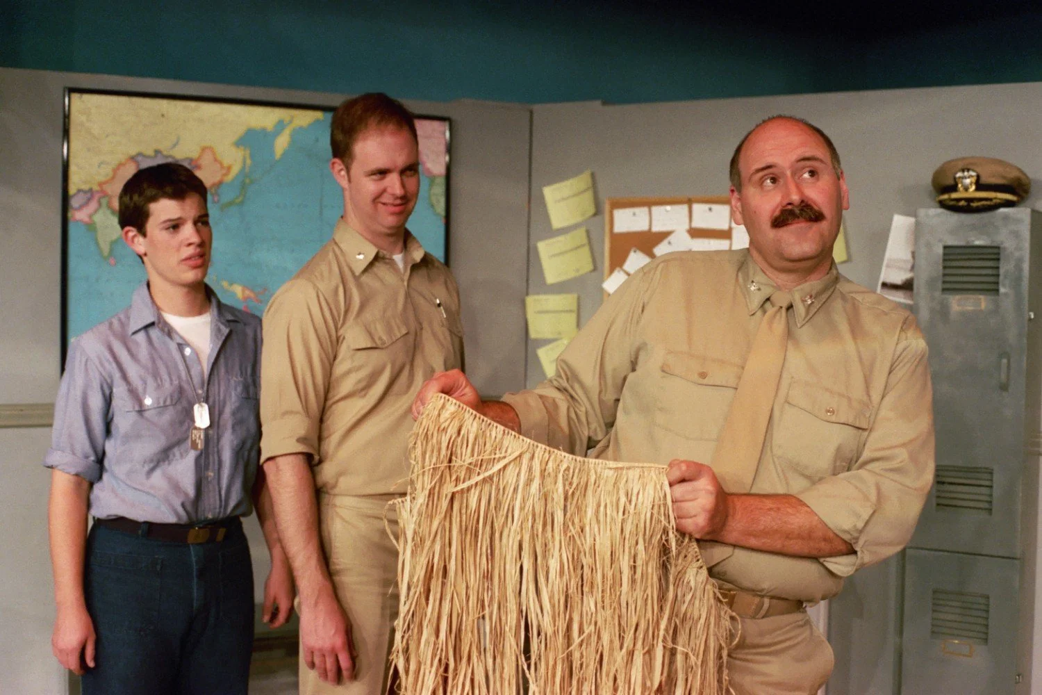 Three men in military uniforms, one holding a grass skirt, in a room with a map and bulletin board.