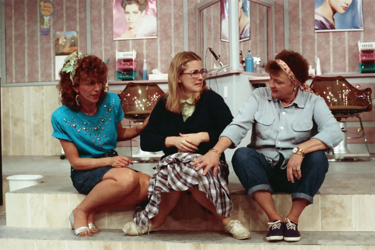 Three women sitting on steps in a hair salon, engaged in conversation. The woman on the left wears a blue shirt with embellishments, the middle woman has glasses and plaid skirt, and the right woman wears a headband and denim shirt. Background includ