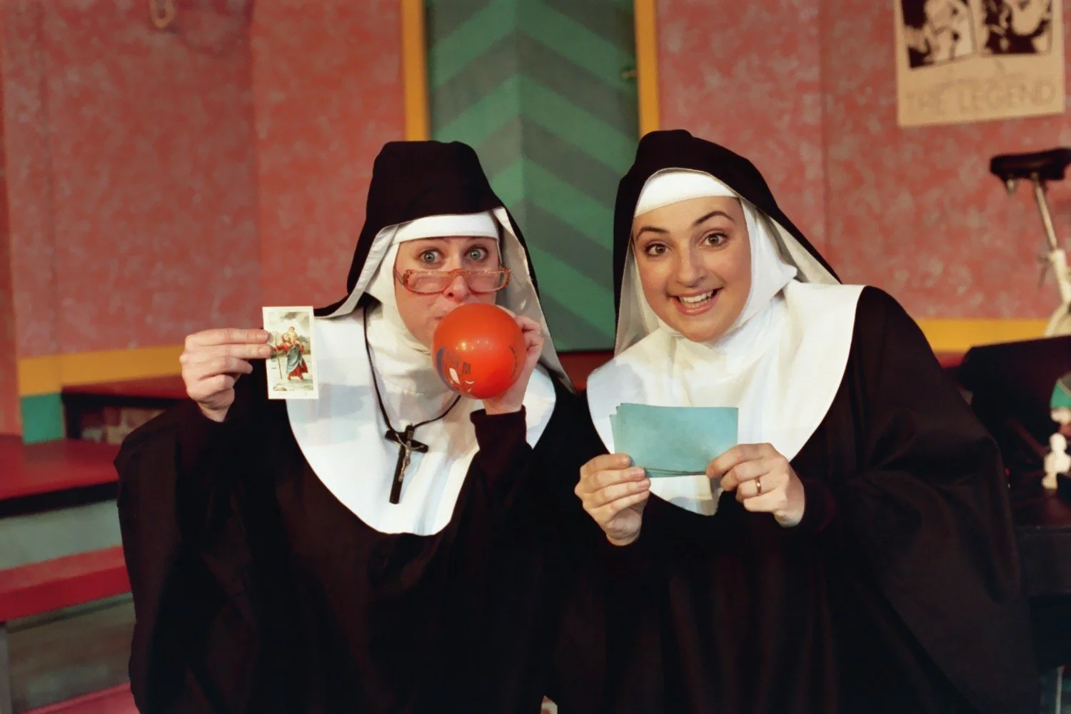 Two women dressed as nuns, one holding a card and red ball, the other holding papers, in a colorful room.