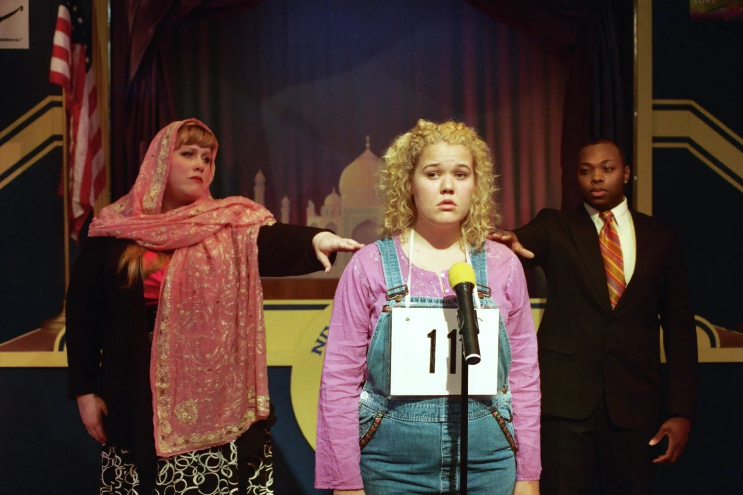 A person wearing denim overalls and a pink shirt stands behind a microphone with a number 11 tag, flanked by two individuals in formal attire against a stage backdrop.