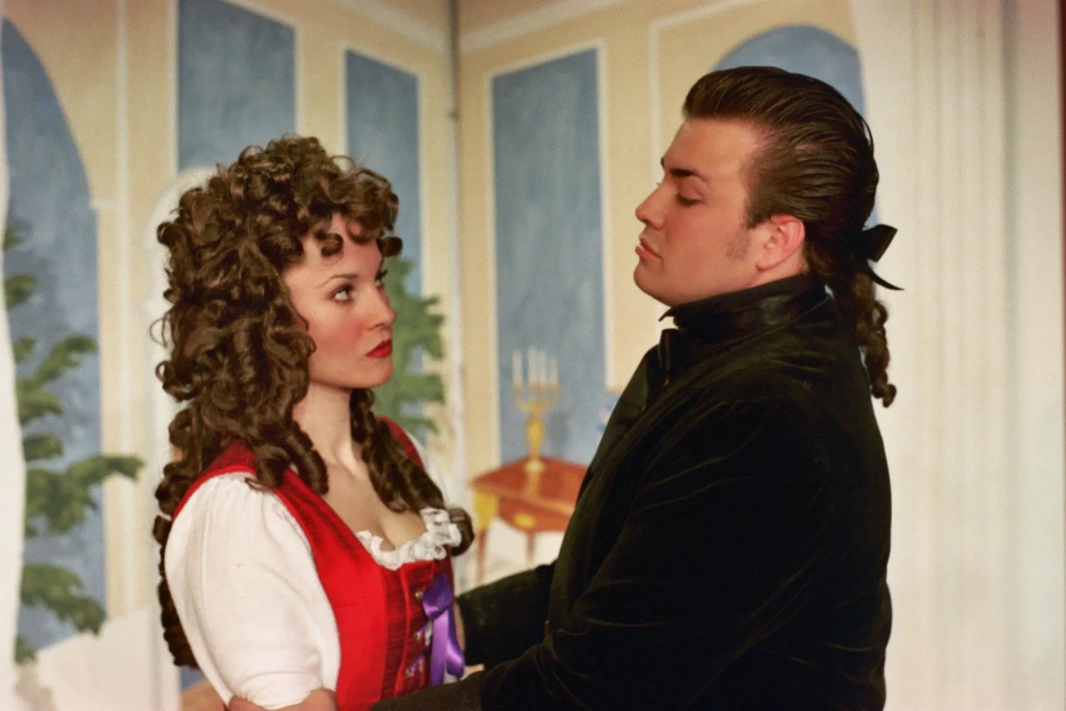 Two people in costume embrace, in a setting with classical decor.