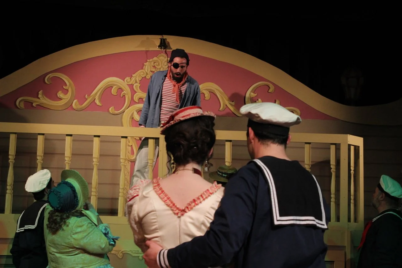 Theatrical performance featuring actors in period costumes on a stage set with a balcony. One actor with a pirate look leans over the balcony, while others in sailor and Victorian-style attire look up.