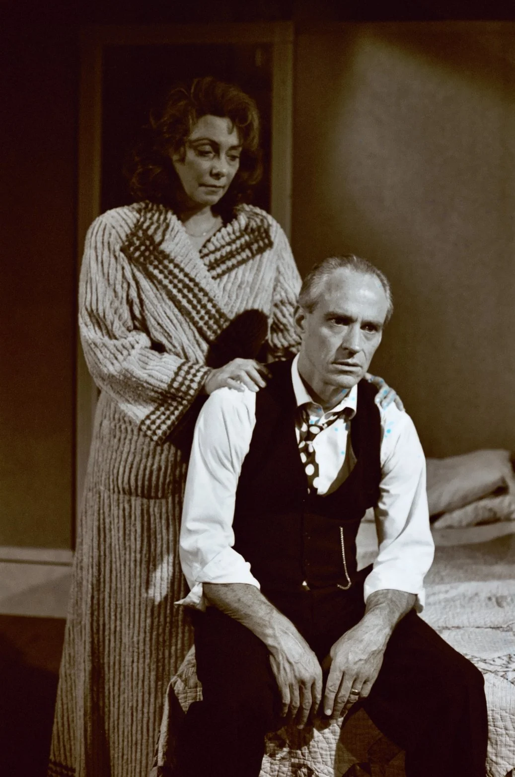 A black-and-white photo of a woman in a robe standing behind a seated man in a vest and polka dot tie, both looking pensive in a domestic setting.