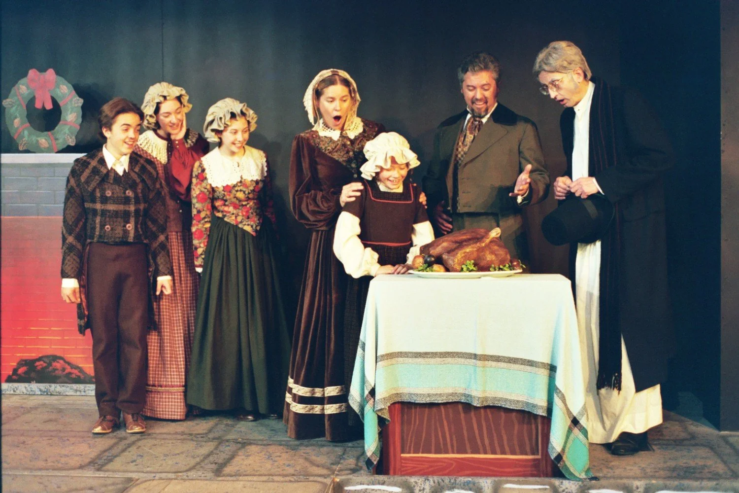 Group of people in Victorian costumes standing around a table with a roast turkey, appearing to be from a theatrical performance.
