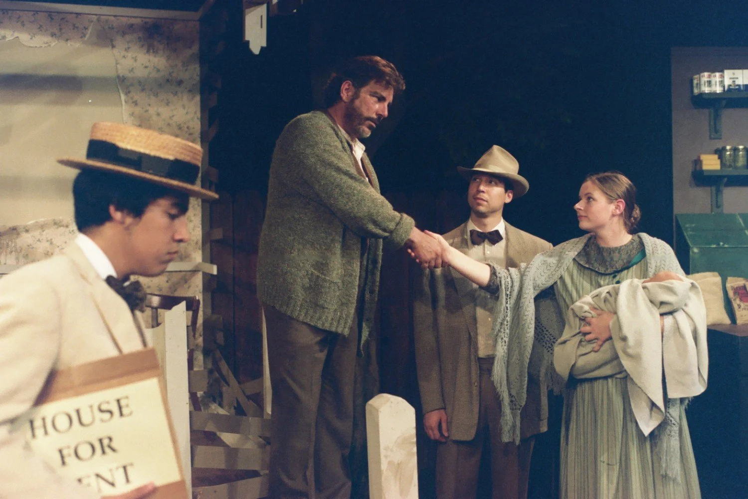 Four actors performing a stage play, with one man holding a "House for Rent" sign, another man and a woman shaking hands while she holds a baby, and a third actor standing between them wearing a suit and hat.