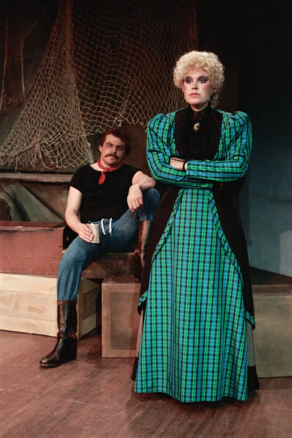 Two actors in a theater set; one is sitting casually, wearing a black shirt and red neckerchief, and the other is standing in a green plaid dress, looking serious.