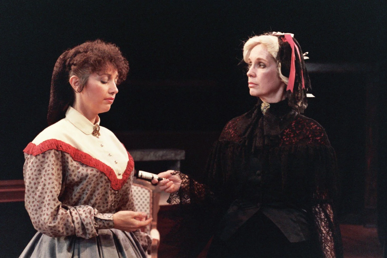 Two women in period costumes, one in a light colored dress with red trim, the other in a dark dress with lace, exchanging an item.