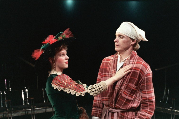 Two actors on stage in Victorian costumes, with one wearing a green dress with lace and a hat, and the other in a red plaid robe and white headscarf.