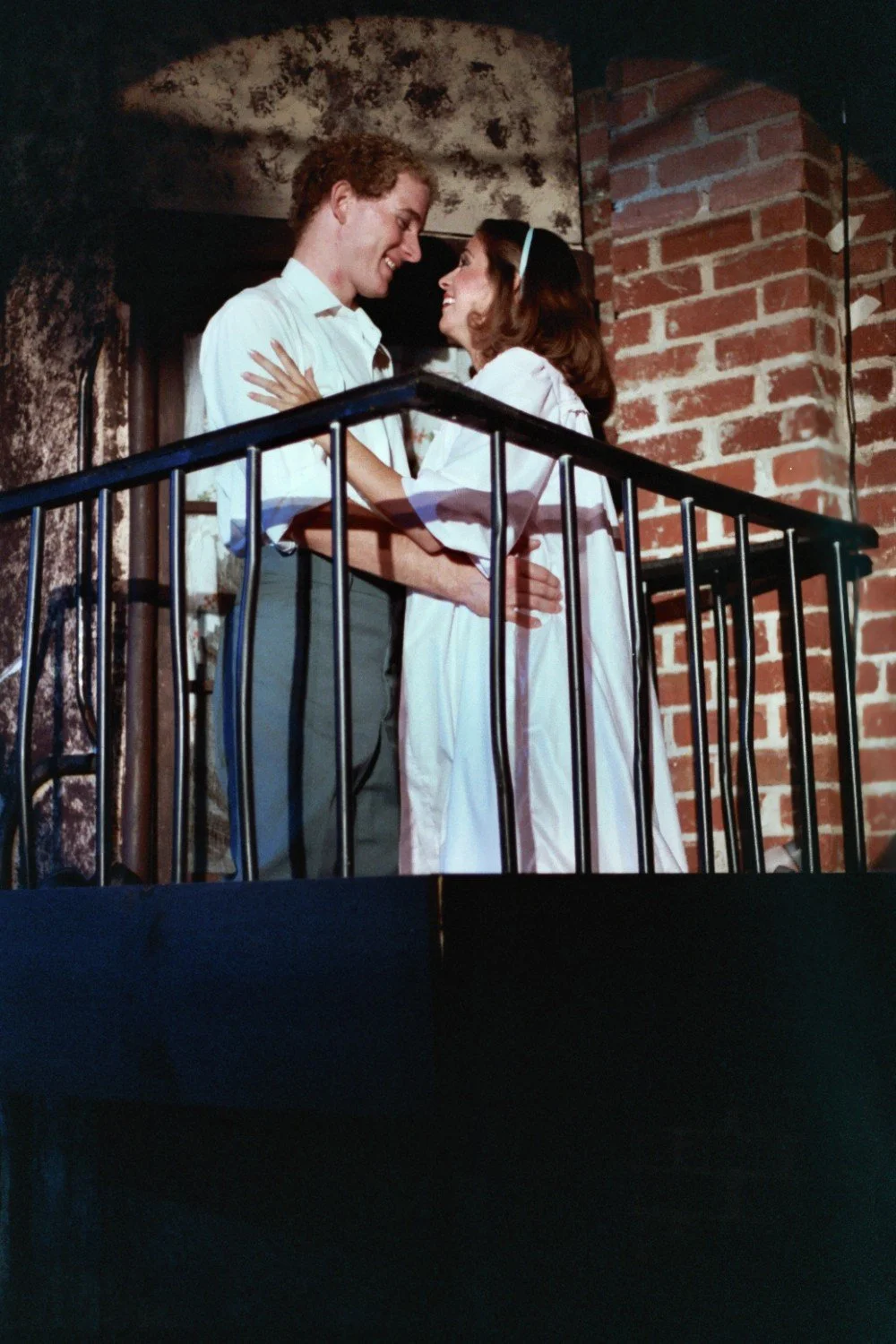 A man and woman embracing on a balcony with brick and textured walls, smiling at each other.