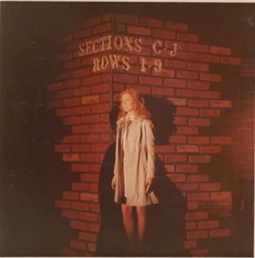 Woman in a coat standing by a brick wall with white text "Sections C-J Rows 1-9."