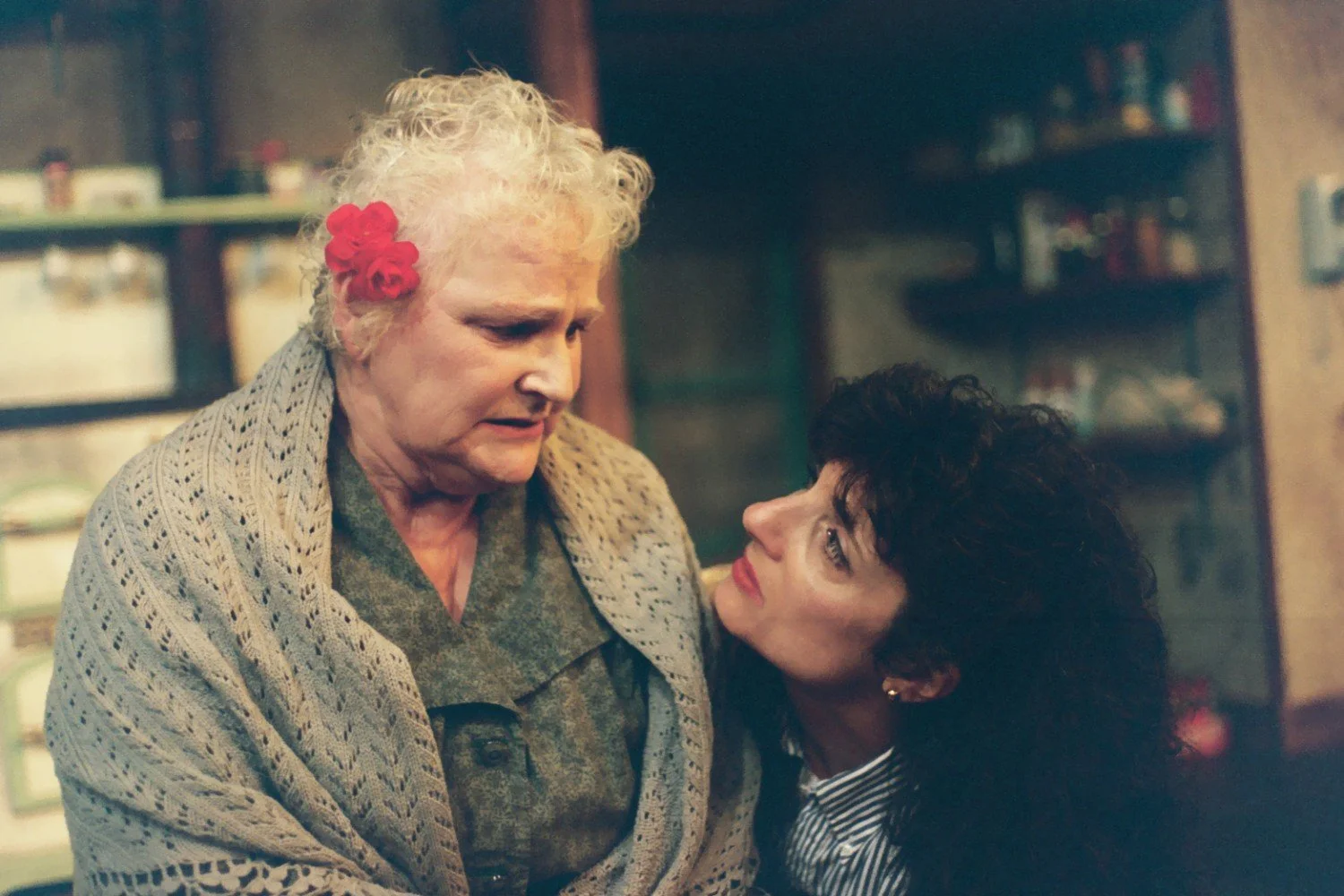 An elderly woman with curly white hair and a red flower tucked beside her ear, wrapped in a shawl, looking downward with a concerned expression. A younger woman with dark hair gazes up at her with an attentive and caring expression, set in a cozy, di