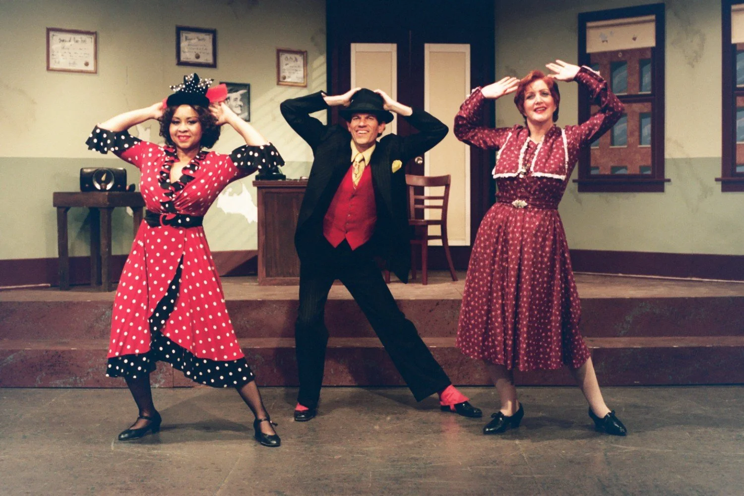Three people in vintage-style outfits performing a dance on stage, with a theater set depicting an old-fashioned room in the background.