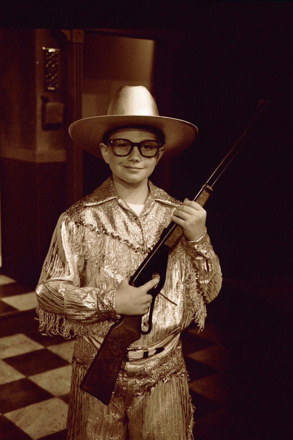 Boy in cowboy costume holding a toy rifle, wearing glasses and a wide-brimmed hat.
