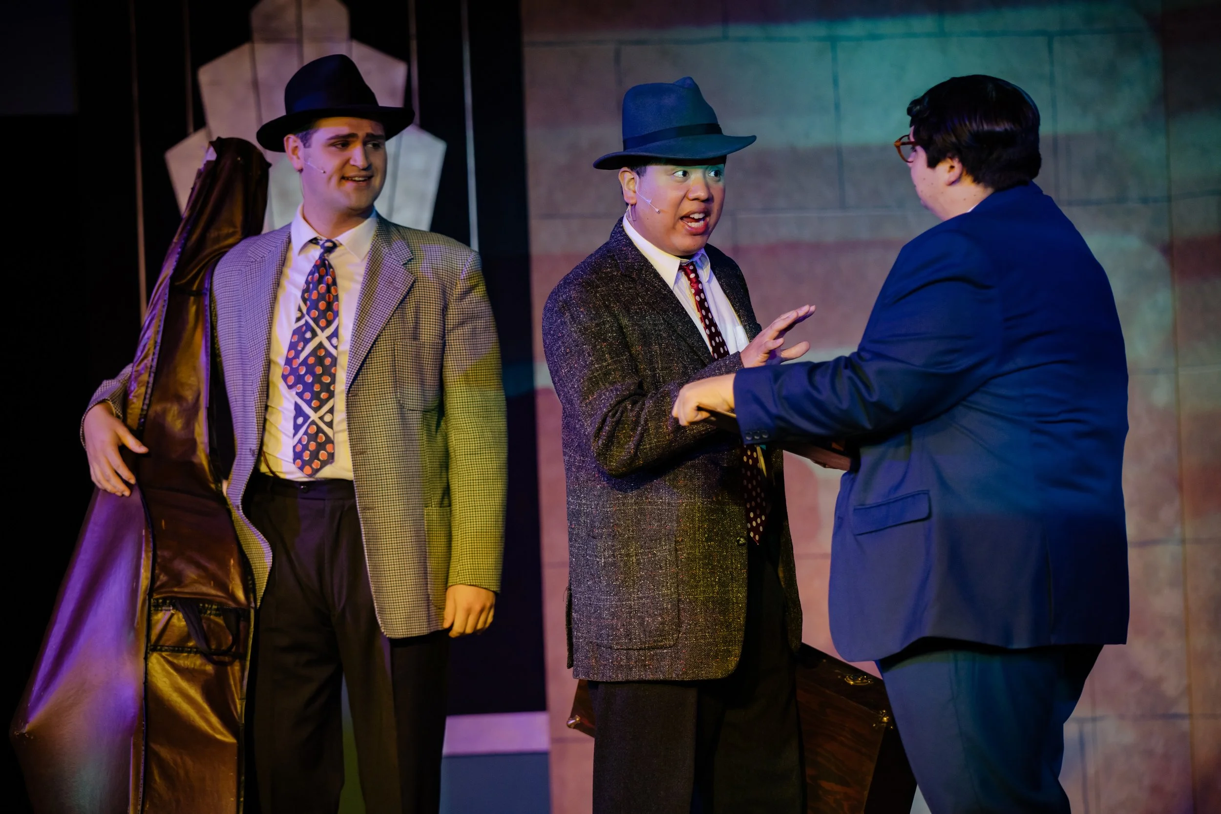Three men in vintage suits and hats on stage, one holding a double bass case, engaged in conversation during a theatrical performance.