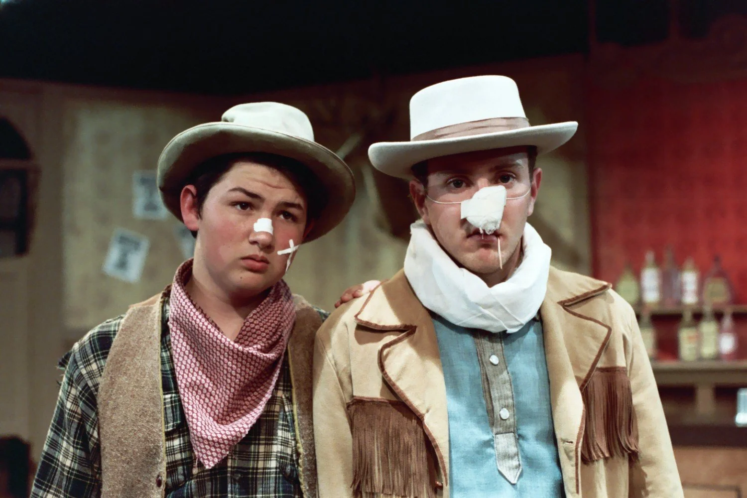 Two men in Western costumes with bandages on their noses, wearing hats and standing indoors.
