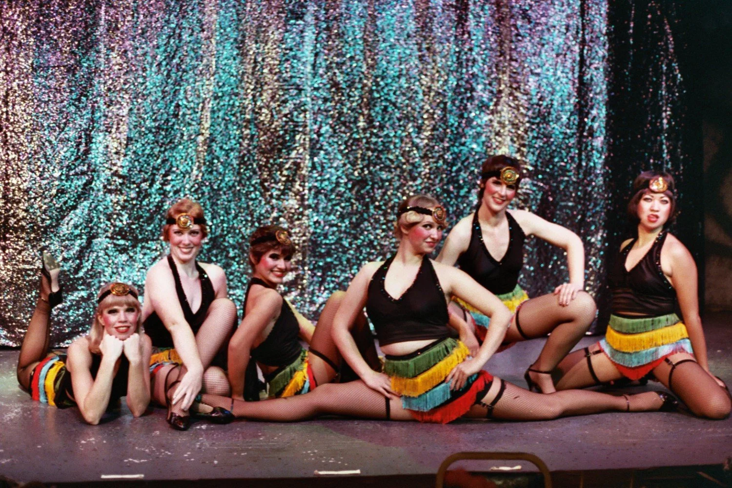 Six performers in colorful fringe skirts and black tops, posing on a stage with a glittery backdrop, resembling a dance or theater performance.