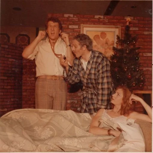 Three people in a living room setting: a man wearing a robe using a stethoscope on another man, who is making a surprised expression, and a woman lying on a couch, looking up at them. There is a decorated Christmas tree in the background.