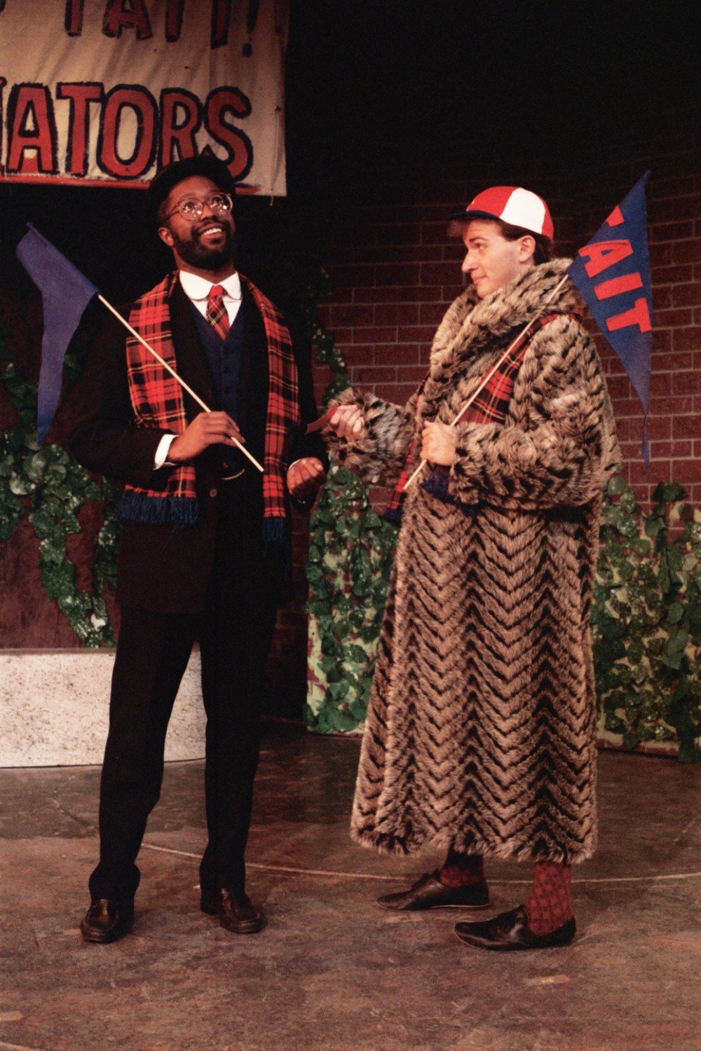 Two people in colorful, theatrical costumes holding flags and talking in front of a brick wall with greenery.