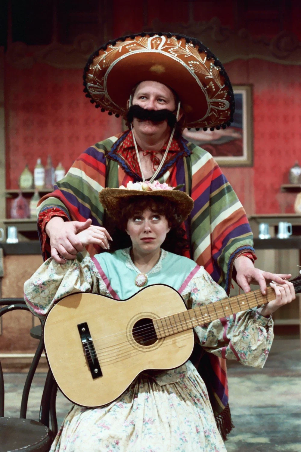 Two people in costume, one wearing a large sombrero and colorful poncho with a fake mustache, standing behind the other who is seated and holding a guitar. The seated person wears a floral dress and straw hat, looking upward.