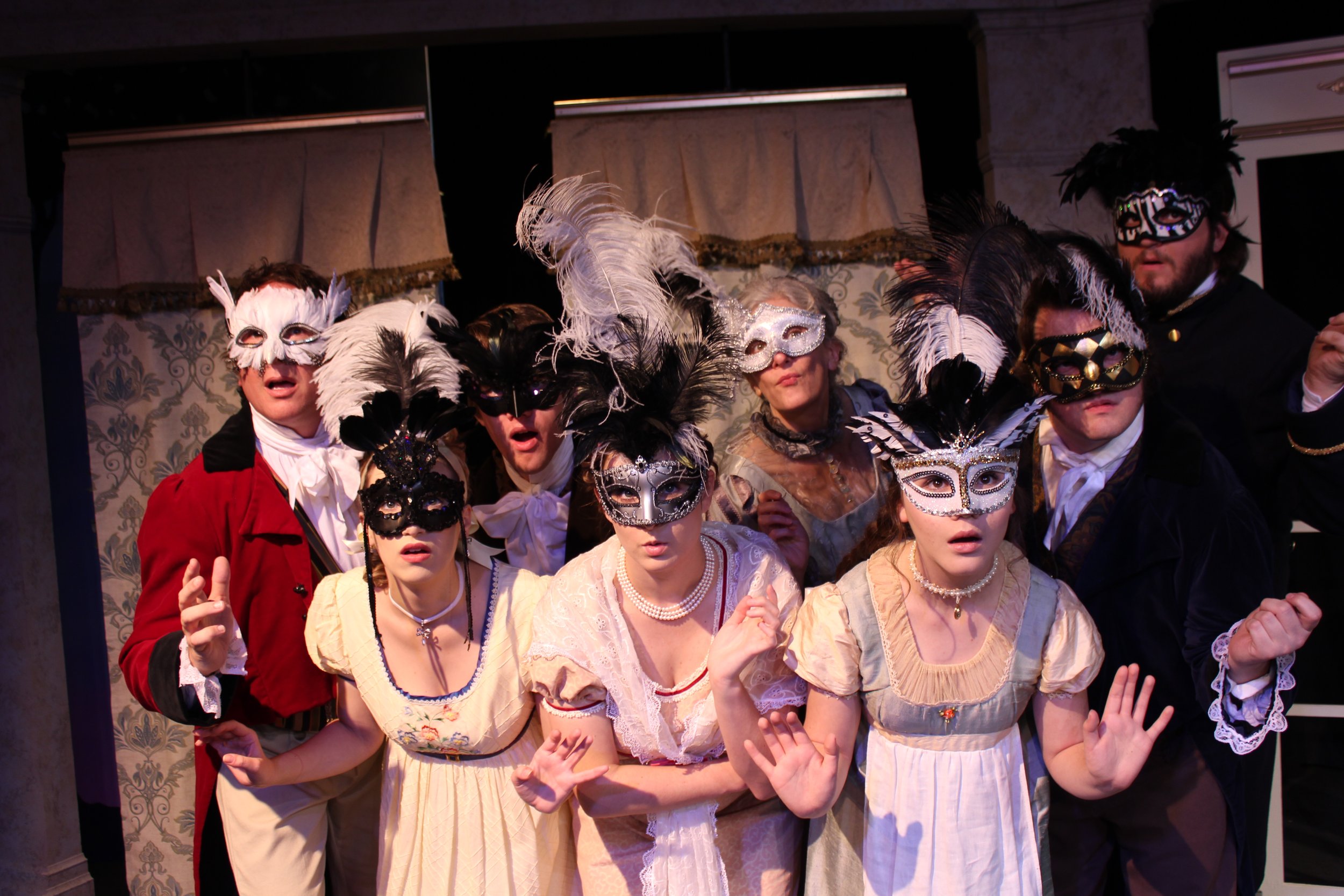 Group of people dressed in formal historical attire with decorative masks, gathered closely, in a theater setting.