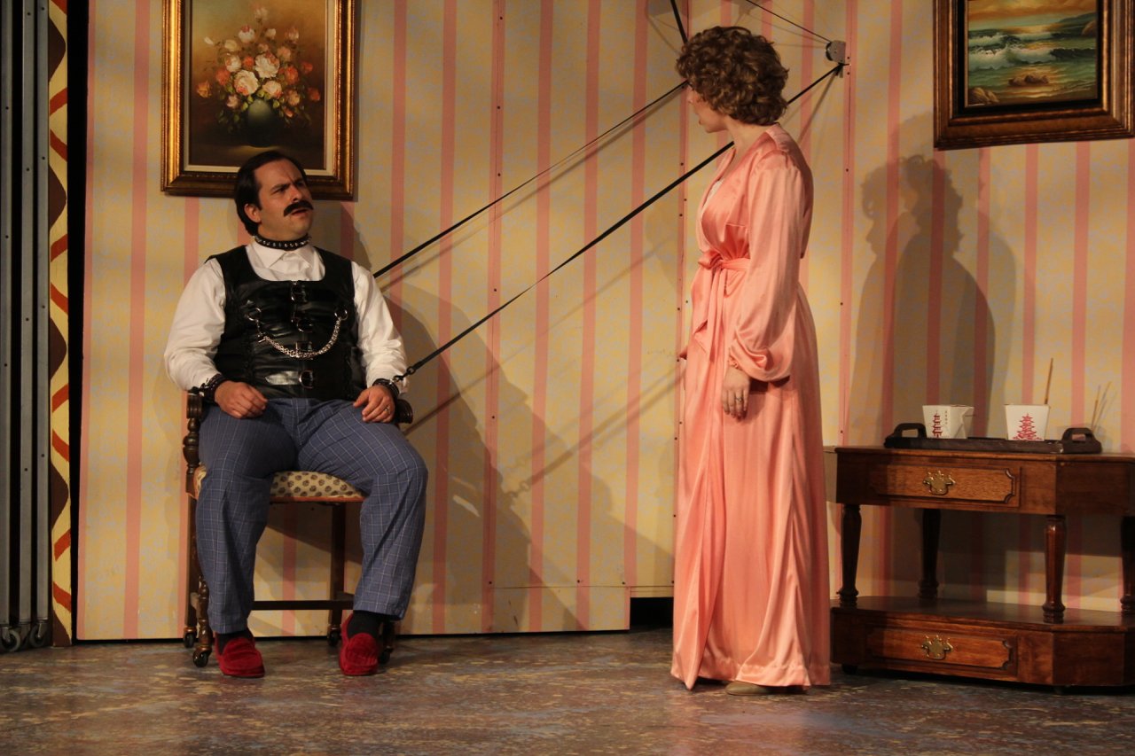 Two actors on stage in a play. One is sitting on a chair, wearing a leather vest and chains, and the other is standing in a pink robe. The room has striped wallpaper and framed paintings on the walls. A wooden table with Chinese takeout boxes is visi
