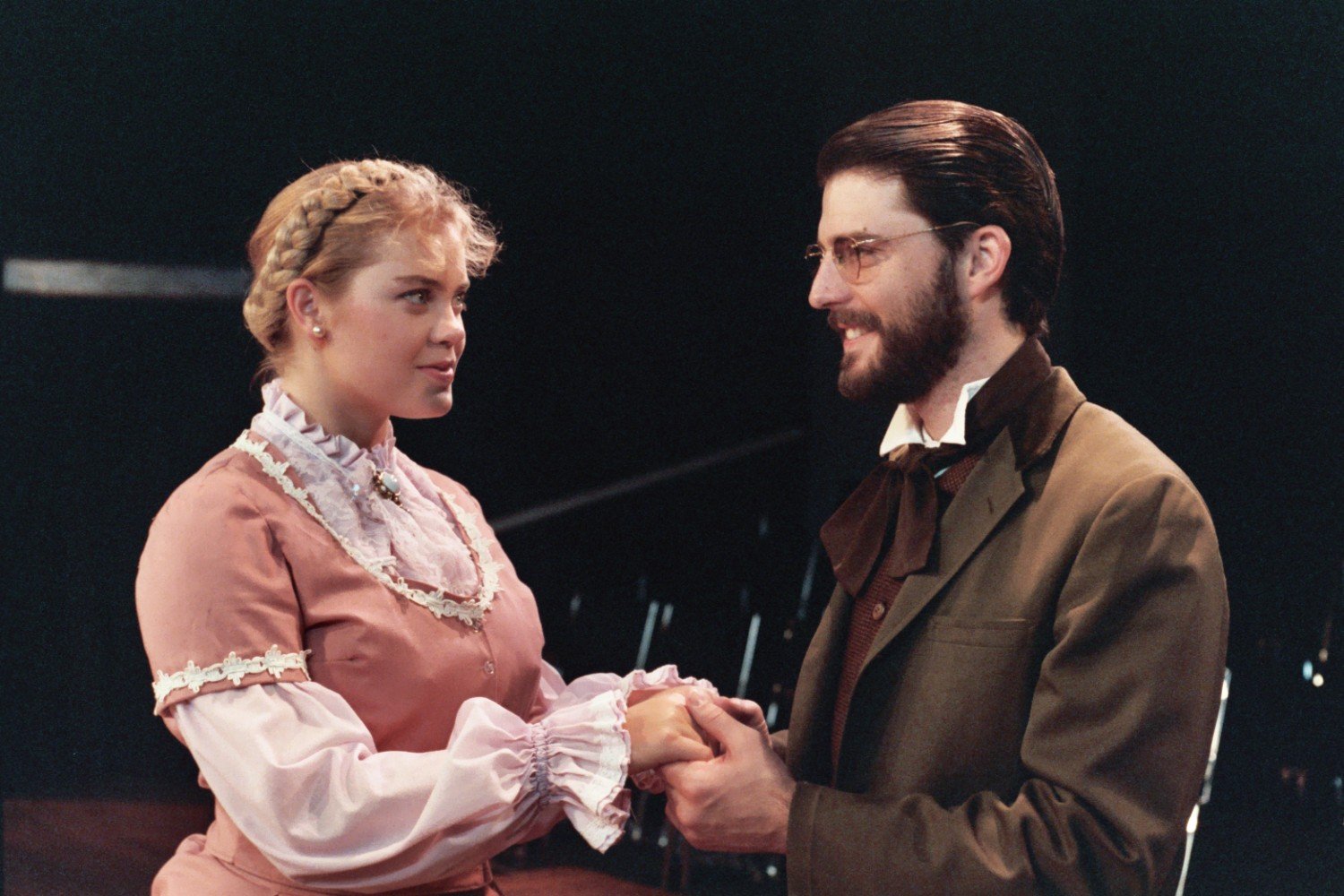 Two people in vintage attire, holding hands and smiling in a theatrical setting.