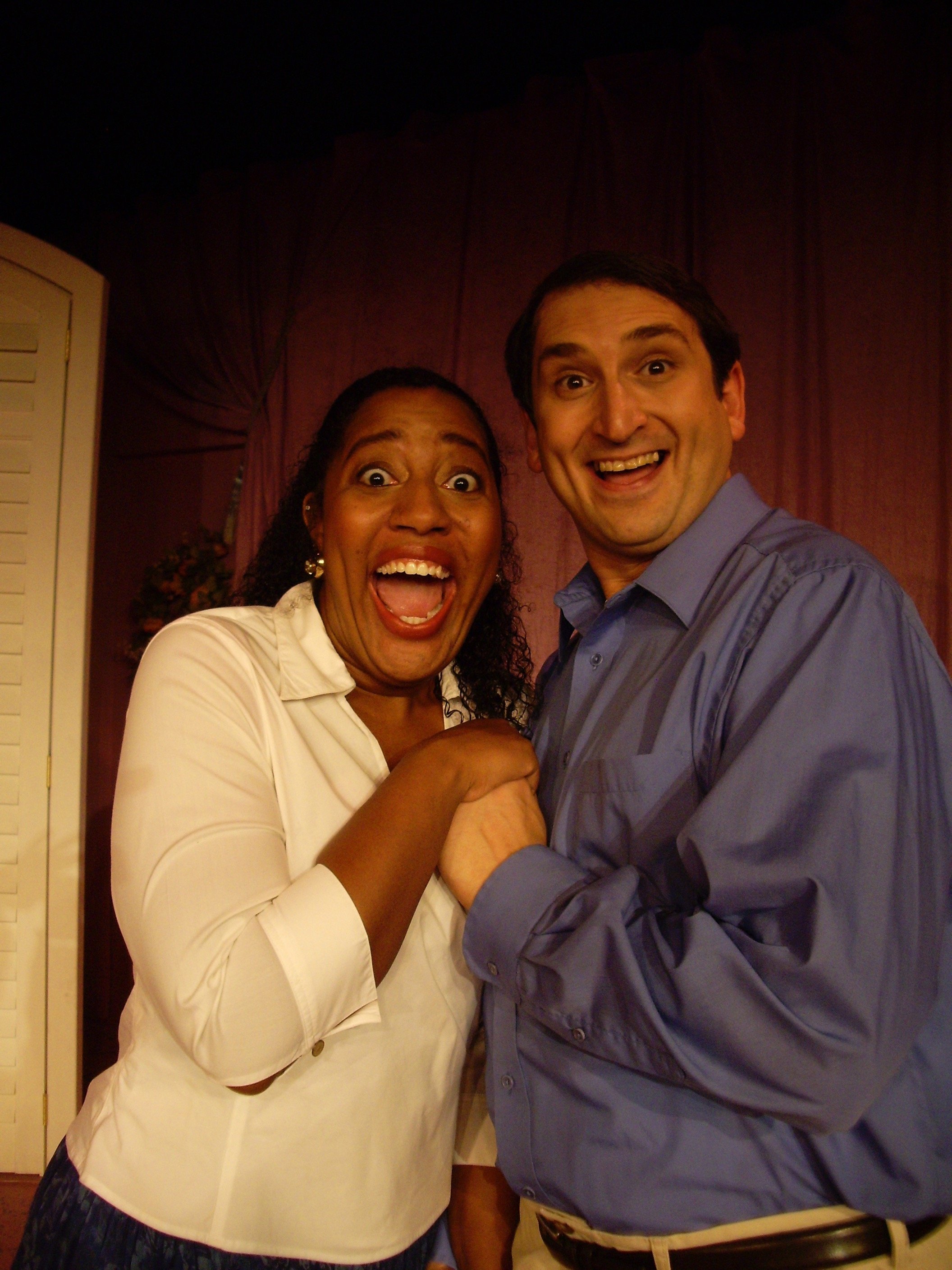 Two people smiling broadly, holding hands, with a curtain backdrop.