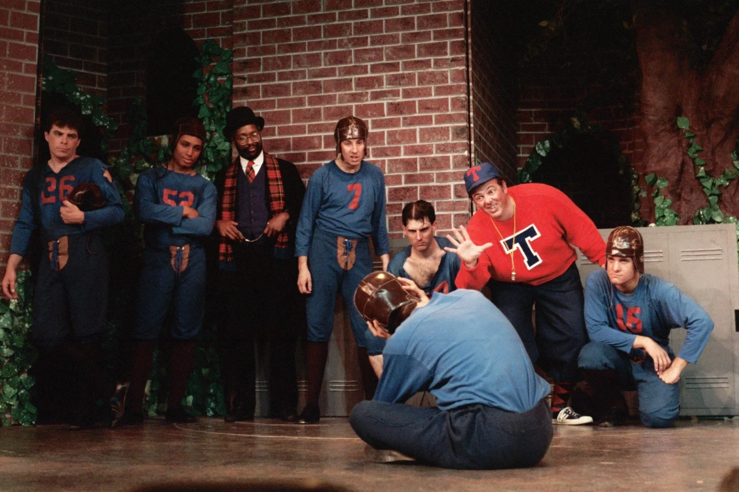 Group of men in old-fashioned football uniforms and a coach in a red sweater with a 'T' emblem, standing in front of a brick wall and lockers, some wearing leather helmets.