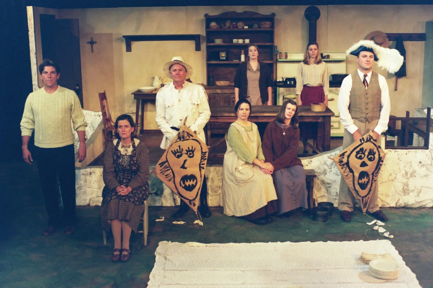 A group of people in vintage clothing pose in a set resembling a rustic interior. Two men at the ends hold masks. The others sit or stand in front of a kitchen area with shelves and cookware. The scene appears theatrical.