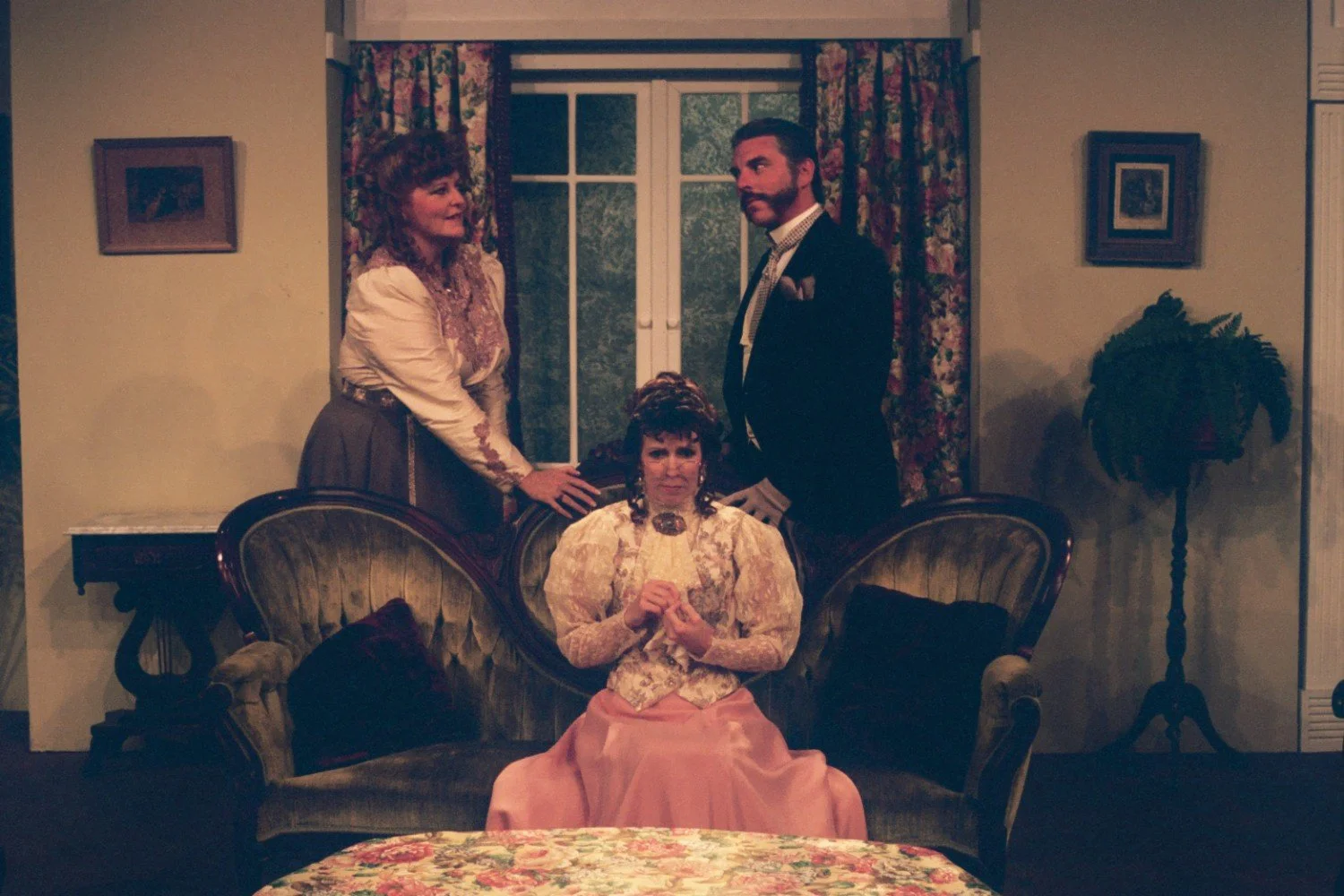 Three people in Victorian-era clothing, with a woman sitting on a vintage couch and a man and woman standing behind her. The room is decorated with floral curtains, a potted plant, and framed pictures on the wall.