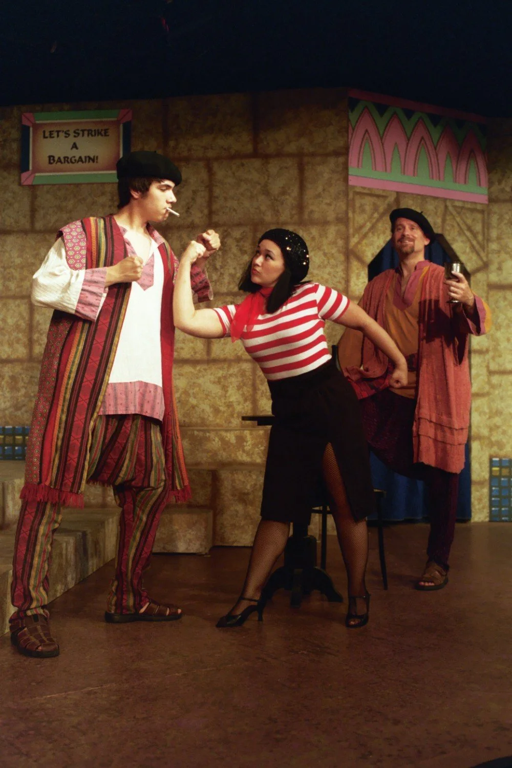 Theatrical performance with three actors in colorful costumes; one actor flexes muscles, another holds a drink, and a sign in the background reads "Let's strike a bargain."