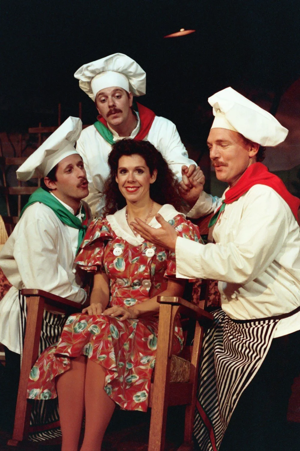 Group of people in costumes performing a comedic scene, with a woman seated and three men dressed as chefs.