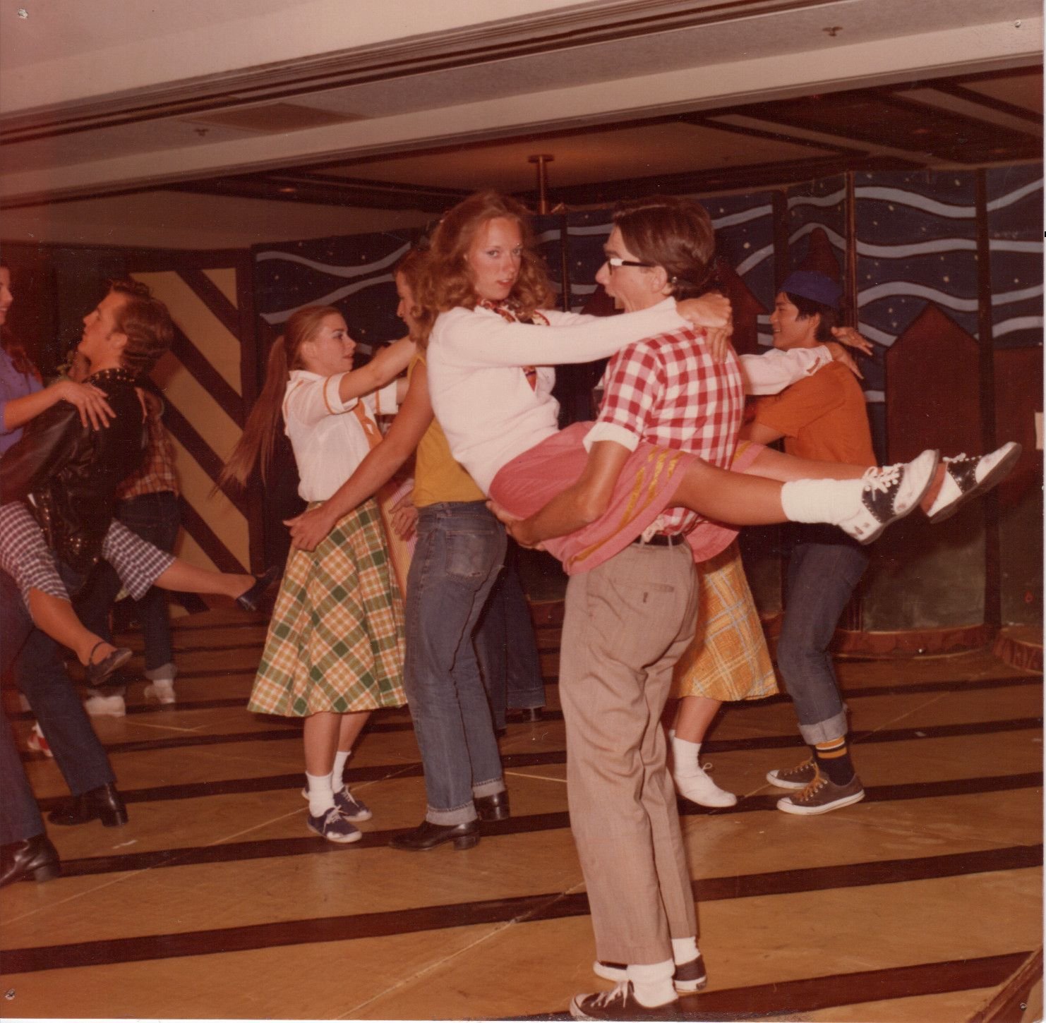 Group of people dancing energetically in a retro-themed setting, wearing vintage attire, with one person lifting another in a playful pose.