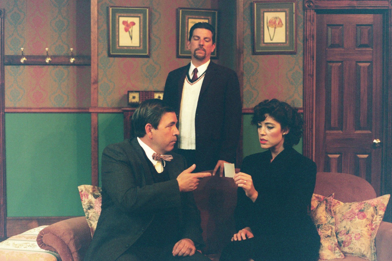 Three people in a vintage room setting; the man seated on the left is gesturing with his hand, while the woman on the right is holding and examining a small card. A third man stands between them in the background. The room has floral patterned wallpa