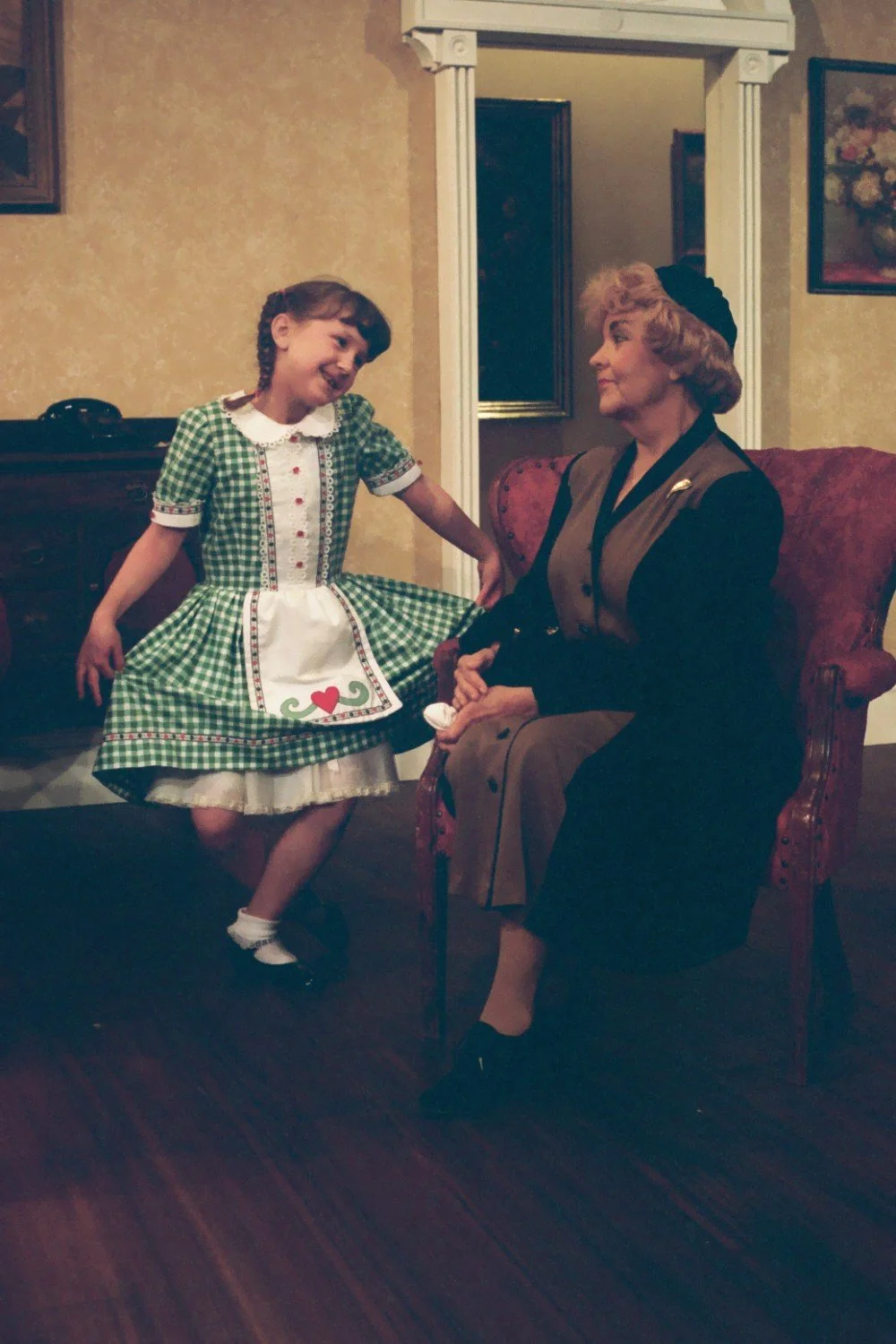 A young girl in a green checkered dress with a white apron and an older woman in a brown and black outfit sitting in a vintage-style room.