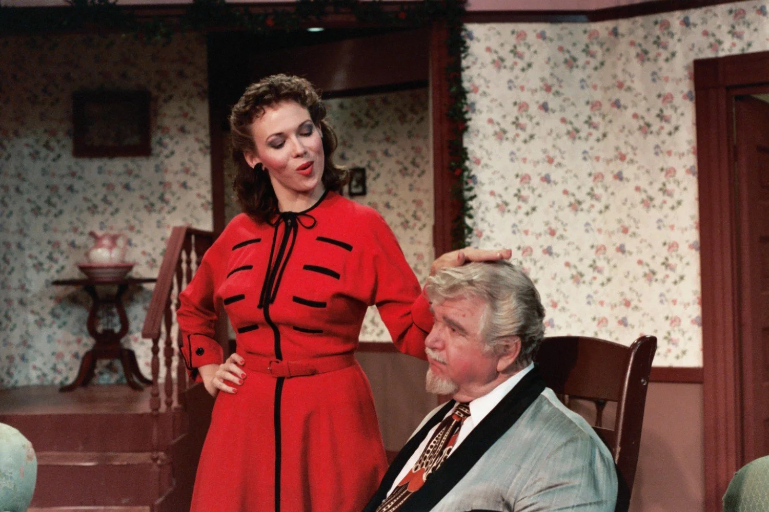 A woman in a red dress touching a seated man's head in a vintage or old-fashioned living room setting with floral wallpaper and wooden furniture.