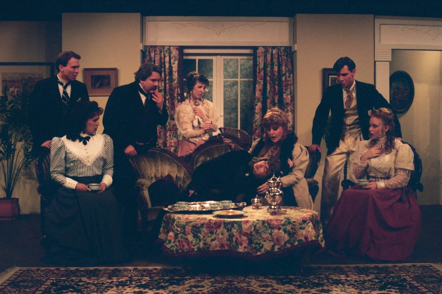 Victorian-era play scene with actors in period costumes, gathered around a fainting person on a couch, in a vintage living room setting with floral curtains and a decorated table.