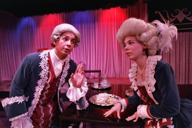 Two people in period costumes performing on stage, wearing 18th-century-style wigs and clothing, with a backdrop of red and blue curtains.