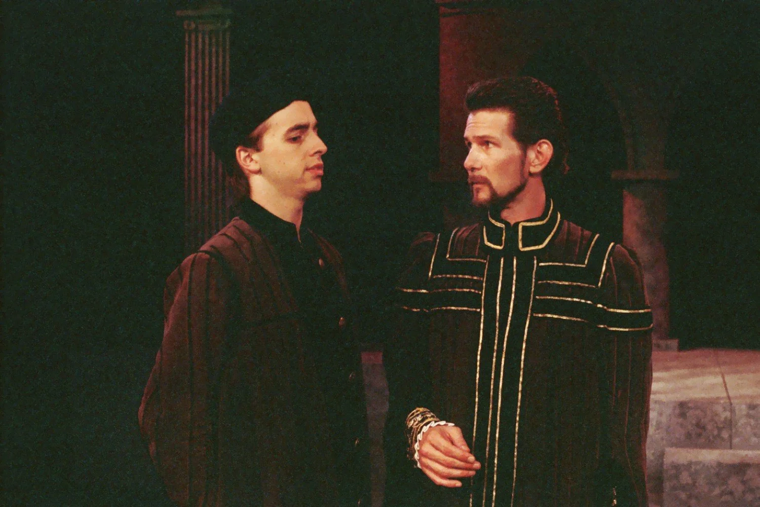 Two actors in period costumes on stage, engaged in conversation, with a dark background and architectural elements.