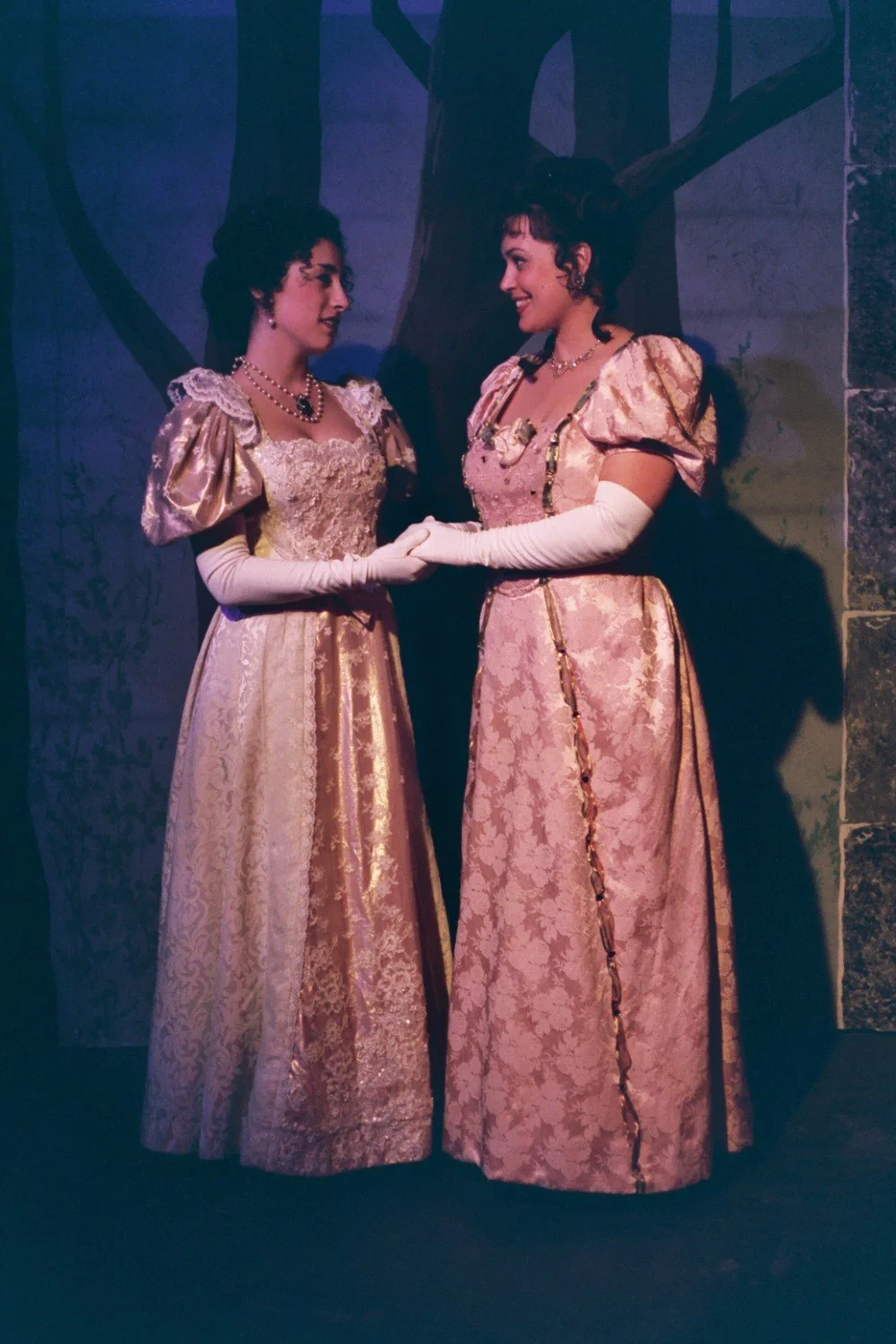 Two women in Regency-era dresses holding hands on a dimly lit stage with a painted backdrop.