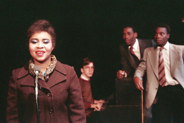 Woman singing with a microphone on stage; three men in the background, including one playing the piano.