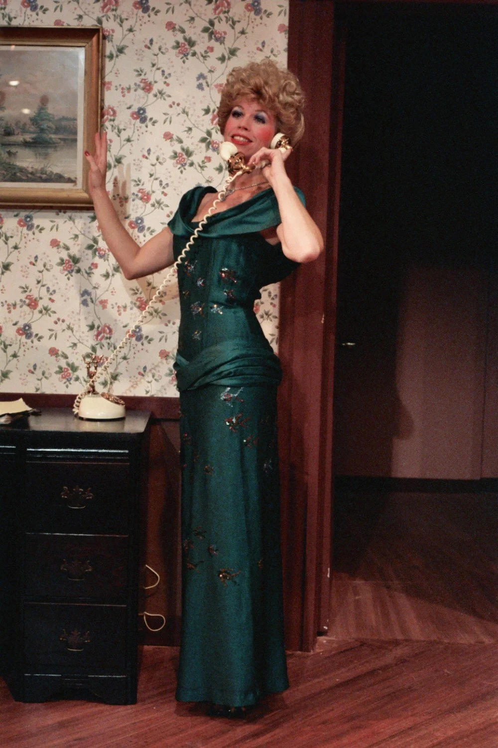 Person in a green evening gown talking on a vintage telephone in a room with floral wallpaper and a framed painting.
