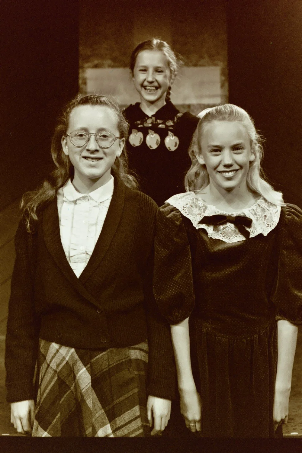 Three girls in vintage-style clothing smiling; one in the back and two in the front, wearing glasses and dresses with lace and plaid details. Sepia tone.