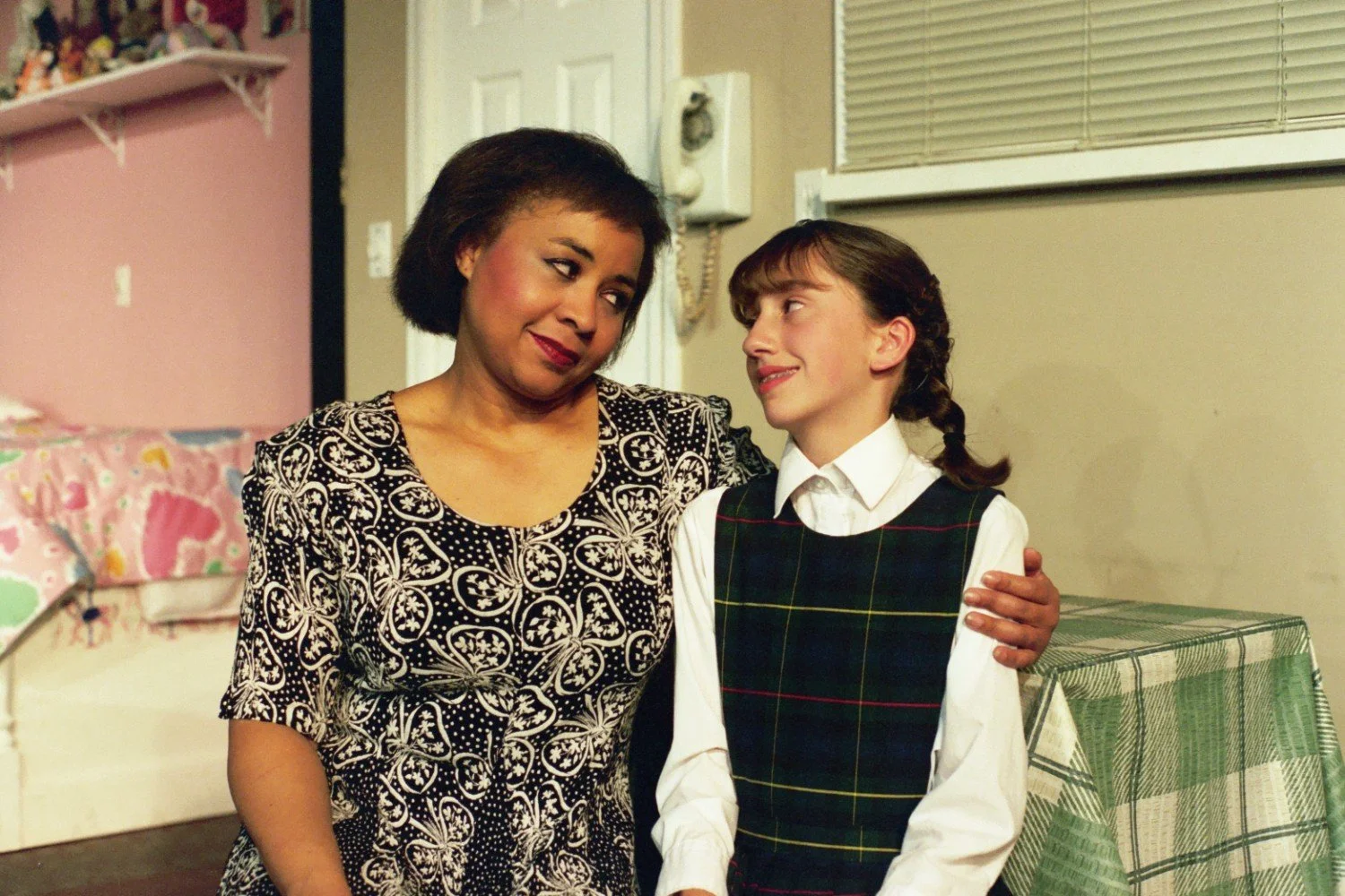 A woman and a young girl smiling at each other. The woman is wearing a black and white patterned dress, and the girl is in a plaid dress with a white shirt. They are in a room with a pink wall, bed, and a shelf of toys visible in the background.