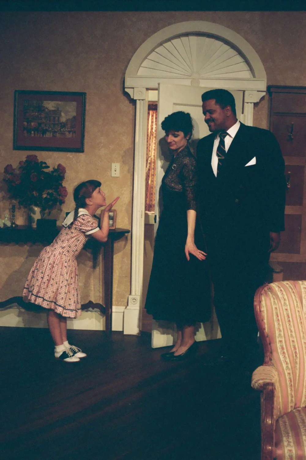 A young girl in a plaid dress poses with two adults in formal attire in a vintage-style room. The girl is leaning over a console table with flowers, while the man and woman stand by a doorway. The room has a striped armchair and framed art on the wal